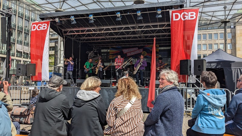 Menschen sitzen vor einer Bühne. Auf Bannern steht "DGB". Auf der Bühne spielt eine Band.