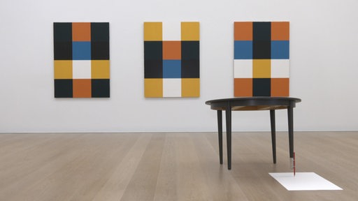 Drei abstrakte Bilder mit quadratischen Motiven hängen in einer Ausstellung hinter einem Tisch