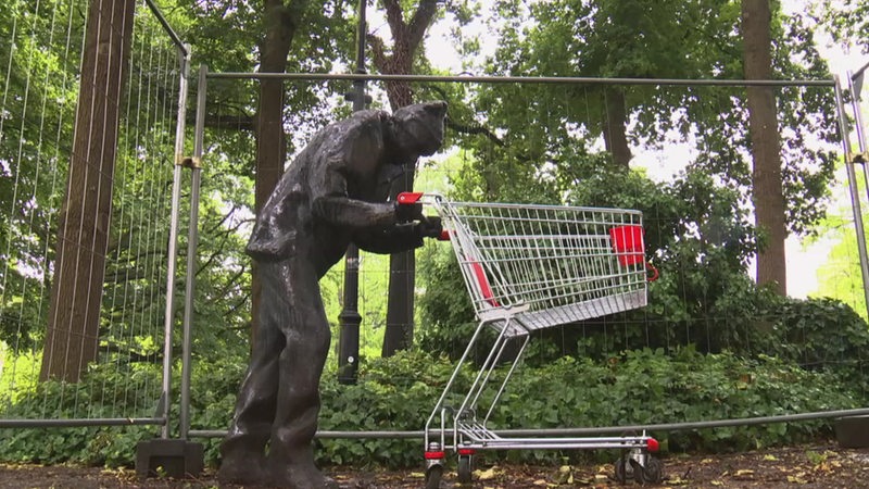 In einem Park steht eine Statue von einem Mann aus Bronze, der einen Einkaufswagen schiebt.