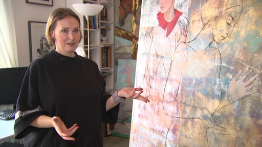 Eine Frau steht vor einem Gemälde, welches eine weitere Frau zeigt, und gestikuliert mit ihren Händen.