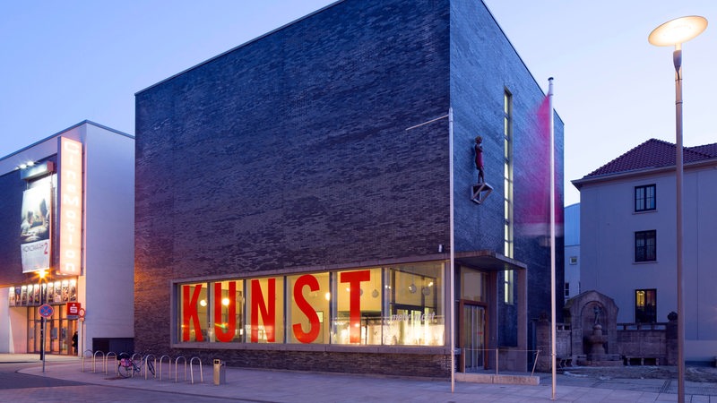 Blick auf ein kastenförmiges Backsteingebäude. Im Fenster steht in großen Lettern "KUNST".