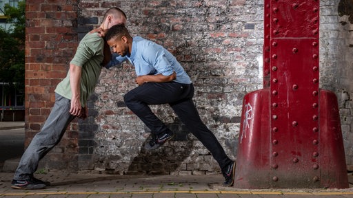 Zwei junge Männer tanzen aneinander gelehnt vor einer Mauer