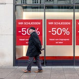 Ein Mann geht über einen Bürgersteig, hinter ihm ist ein leeres Ladenfenster mit Schildern, auf denen "Wir schließen!" steht.