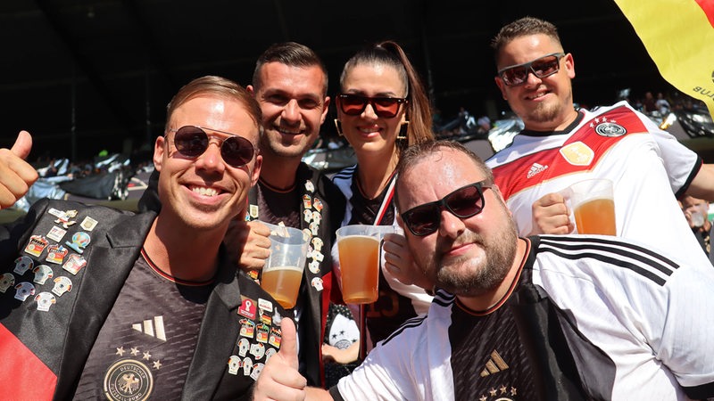 Deutsche Fans stoßen auf der Tribüne des Weser-Stadions vor dem Länderspiel an und schauen gut gelaunt in die Kamera.