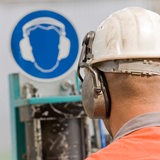 Ein Mann mit Helm und Gehörschutz steht vor einem Gehörschutz-Schild.