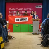 Die Bremer Landessprecherin der Partei Die Linke, Anna Fischer, steht beim Außerordentlichen Landesparteitag ihrer Partei am Rednerpult. Im Hintergrund sitzen drei Menschen. Im Vordergrund sitzen Menschen in mehreren Reihen auf Stühlen.