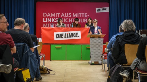 Die Bremer Landessprecherin der Partei Die Linke, Anna Fischer, steht beim Außerordentlichen Landesparteitag ihrer Partei am Rednerpult. Im Hintergrund sitzen drei Menschen. Im Vordergrund sitzen Menschen in mehreren Reihen auf Stühlen.