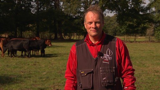 Ein Landwirt steht für ein Interview auf einer Wiese. Im Hintergrund sieht man eine Weide und Rinder.