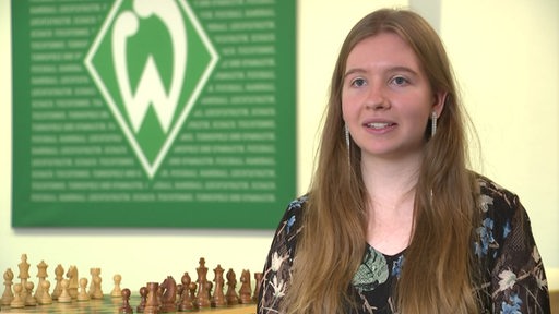 Lara Schulze ist Europameisterin im Schachspielen. Sie sitzt bei einem Interview vor einem Schachspiel und im Hintergrund ist das Logo von ihrem Team Werder Bremen.