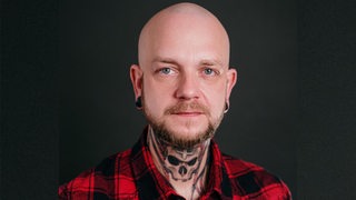 Ein Mann mit Totenkopf-Tattoo am Hals schaut in die Kamera.