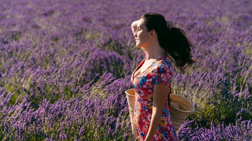 Eine junge Frau steht in einem Lavendelfeld, ihre langen Haare wehen leicht im Wind,.