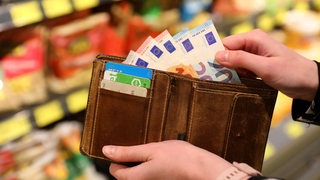 Eine Hand nimmt Geldscheine auf ihrem Geldbeutel. Im Hintergrund sind Supermarktregale unscharf zu erkennen.