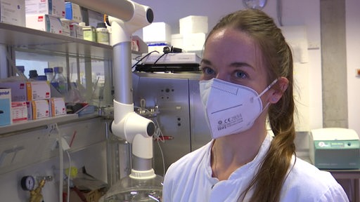 Eine Frau mit Maske und Kittel steht in einem Labor.
