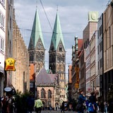 Zahlreiche Menschen sind bei sonnigem Wetter auf der Obernstraße in der Innenstadt unterwegs, während im Hintergrund der historische Bremer Dom zu erkennen ist.