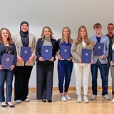 Zwölf Menschen stehen nebeneinander auf einer Bühne und halten jeweils eine blaue Mappe in der Hand.