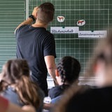 Ein Mann schreibt mit Kreide auf eine große Tafel. In seinem Rücken sitzen Kinder.