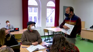 Ein Lehrer hält vor seiner Klasse ein Buch in der Hand in dem er etwas zeigt.