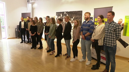 16 Studenten stehen nebeneinander und erhalten ihre Urkunde für ein Lehramtsstipendium in Bremerhaven. 