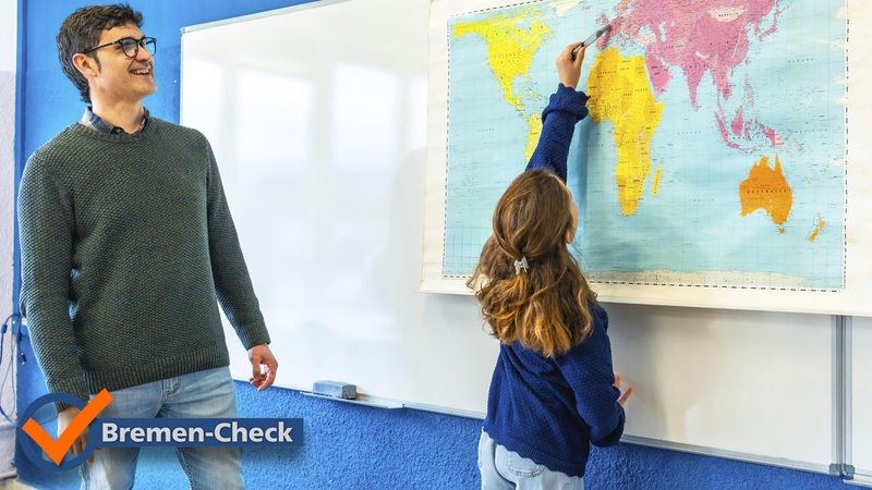 Ein lächelnder Lehrer beobachtet einen Grundschüler, der im Geografieunterricht auf eine Weltkarte zeigt