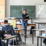 Ein Lehrer lehnt am Lehrerpult vor der Tafel im Klassenraum im Schulunterricht
