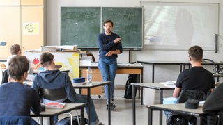 Ein Lehrer lehnt am Lehrerpult vor der Tafel im Klassenraum im Schulunterricht