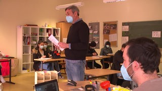 Ein Lehrer, der mitten im Klassenraum steht mit einem Zettel in der Hand und zu den Schülern redet. 