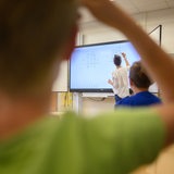 Ein Lehrerin schreibt eine Mathematikaufgabe auf eine digitale Schultafel. Im Vordergrund ist ein Kind zu sehen.