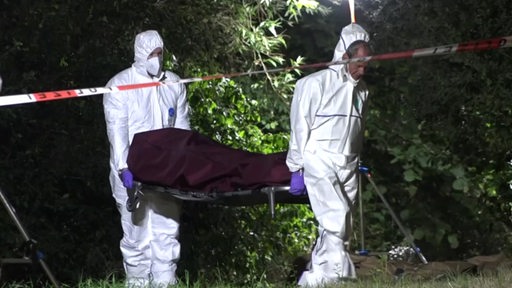 Zwei Männer in weißer Schutzkleidung und mit Masken tragen eine Bahre auf der eine eingewickelte Leiche liegt.