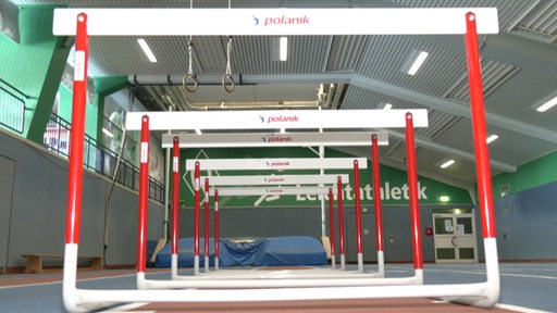 Es sind in einer Sporthalle mehrere hintereinander aufgestellte Hürden zu sehen.