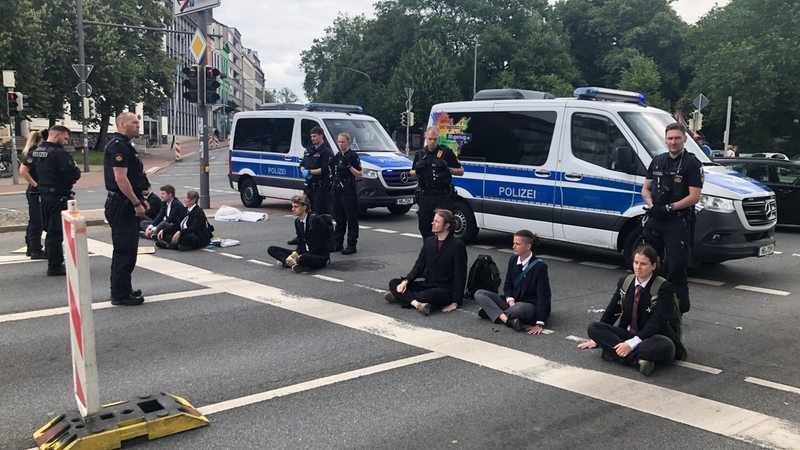 Sieben Menschen sitzen auf einer Straße, Polizisten stehen daneben und dahinter, ebenso Polizeiwagen.