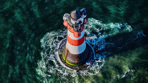 Eine Luftaufnahme zeigt einen rot-weißen Leuchtturm von oben in grün-blauem-Wasser mit weißen Wellen.