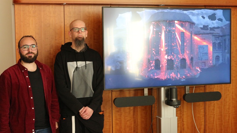 Zwei Männer stehen neben einem Bildschirm.