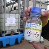 Jemand hält eine Flasche mit weißem, salzartigen Inhalt in die Kamera. Darauf steht: Lithiumhydroxid-Monohydrat.