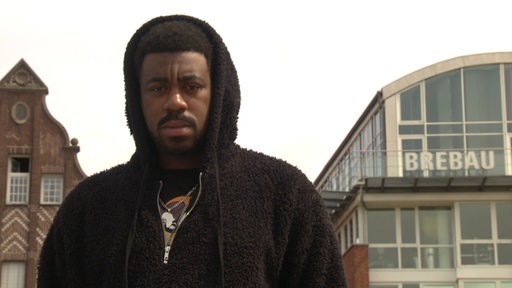 Ein junger schwarzer Mann, Livingsoul Dupe Igunbor, steht vor dem Brebau-Gebäude an der Schlachte in Bremen.