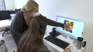 Eine Neuropsychologin steht neben einer Patientin mit Long-Covid und zeigt ihr bei einem Computerspiel zum Gehirntraining etwas auf dem Bildschirm.