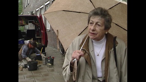 Ein älteres Foto mit einer Frau mit Regenschirm. Im Hintergrund verlegt ein Mann Stolpersteine