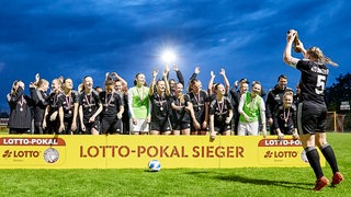 Eine Frau reckt einen Pokal in die Höhe, sie hat der Kamera den Rücken zugewandt. Ihre Mannschaftskolleginnen jubeln ihr zu und haben sich in Richtung Kamera gedreht.