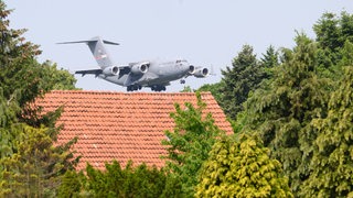 Ein Militärflugzeug fliegt über einem Dach zwischen Bäumen.