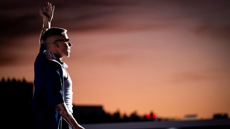 Der Rapper Macklemore steht auf einer Bühne und hebt den linken Arm.
