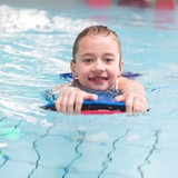 Ein Mädchen im Schwimmbad klammert sich an ein Schwimmbrett und schwimmt in Richtung Kamera. 