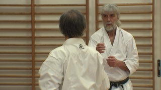 Zwei Männer stehen sich gegenüber. Sie haben eine Kampfpose eingenommen und tragen Karate-Anzüge.