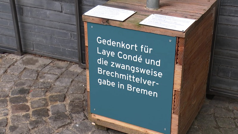 Ein Holzsockel ist zu sehen. Er trägt die Aufschrift: "Gedenkort für Laye Condé und die zwangsweise Brechmittelvergabe in Bremen".