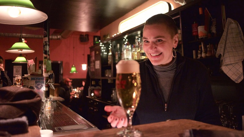 Junge kurzhaarige Frau hinter einer Theke präsentiert ein Bier