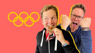 Ein Mann beißt in eine Goldmedaille, dahinter ein jubelnder Mann, beide vor einem roten Hintergrund mit dem Olympia-Logo aus fünf Kreisen.