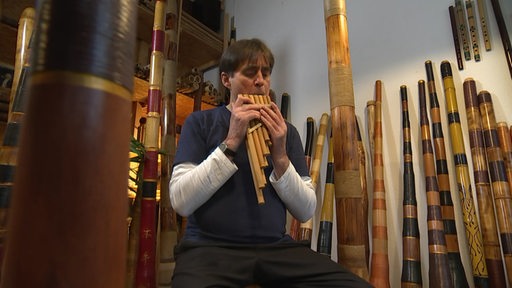 Ein Mann, der eine Bambusflöte spielt und umgeben ist von Didjeridoos.