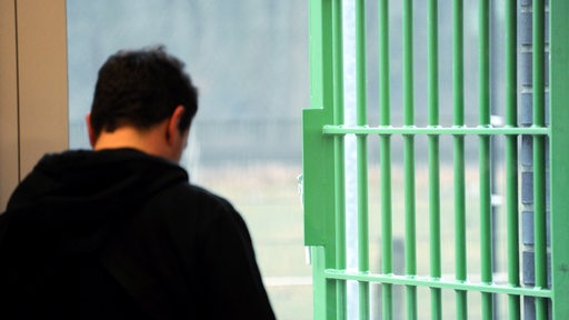 Ein Mann steht in einem Ganag, im Hintergrund ein Fenster und eine Gitterstäbe-Tür