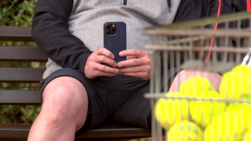 Ein Mann sitzt hinter einem Korb mit Tennisbällen und hält ein Handy in der Hand.