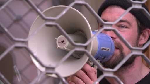 Ein Mann hält einen Lautsprecher vor seinem Mund. Er steht hinter einem Zaun.