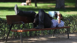 Ein Mann in Hemd und Hosen liegt auf einer Parkbank und liest eine Zeitung.