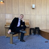 Generalmusikdirektor Marco Comin sitzt lachend auf einem Stuhl vor einem Klavier